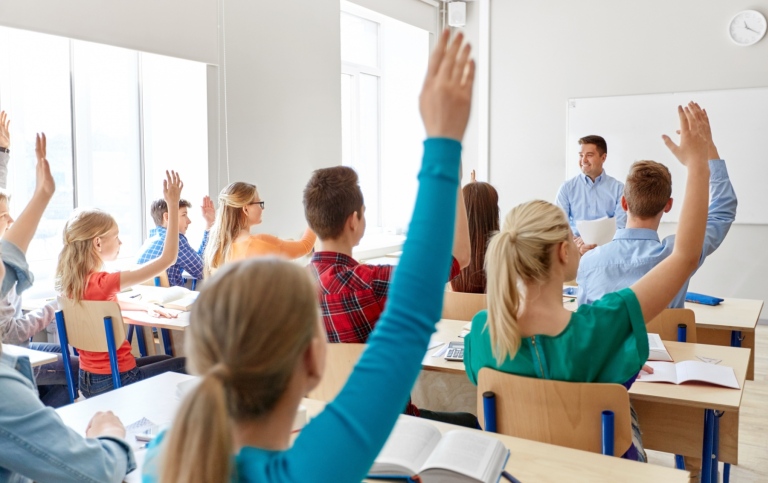 Elever sitter i ett klassrum och räcker upp handen, en manlig lärare ser glad ut